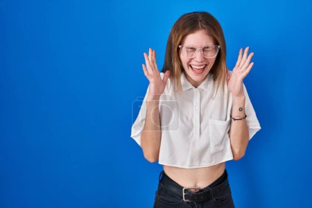 Téléchargez les photos : Beautiful woman standing over blue background celebrating mad and crazy for success with arms raised and closed eyes screaming excited. winner concept - en image libre de droit