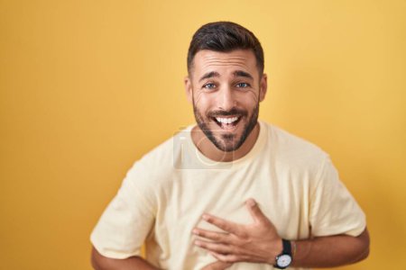 Foto de Hombre hispano guapo de pie sobre fondo amarillo sonriendo y riendo fuerte en voz alta porque divertido chiste loco con las manos en el cuerpo. - Imagen libre de derechos