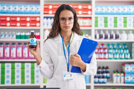 Téléchargez les photos : Young hispanic woman working at pharmacy drugstore holding syrup skeptic and nervous, frowning upset because of problem. negative person. - en image libre de droit