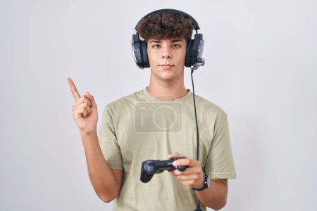 Photo for Hispanic teenager playing video game holding controller with a big smile on face, pointing with hand finger to the side looking at the camera. - Royalty Free Image