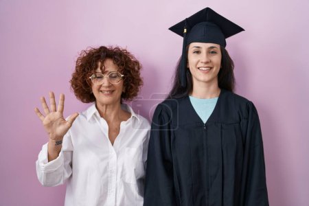 Foto de Madre e hija hispanas con gorra de graduación y bata de ceremonia mostrando y señalando con los dedos número cinco mientras sonríen confiadas y felices. - Imagen libre de derechos