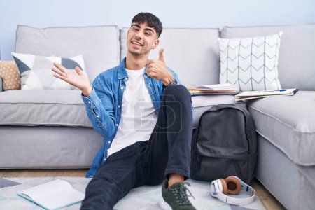 Foto de Young hispanic man sitting on the floor studying for university showing palm hand and doing ok gesture with thumbs up, smiling happy and cheerful - Imagen libre de derechos
