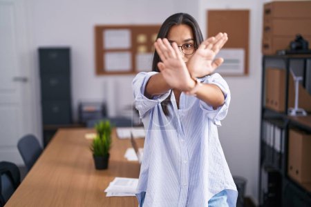 Foto de Young hispanic woman at the office rejection expression crossing arms and palms doing negative sign, angry face - Imagen libre de derechos