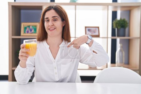 Foto de Brunette woman drinking glass of orange juice looking confident with smile on face, pointing oneself with fingers proud and happy. - Imagen libre de derechos