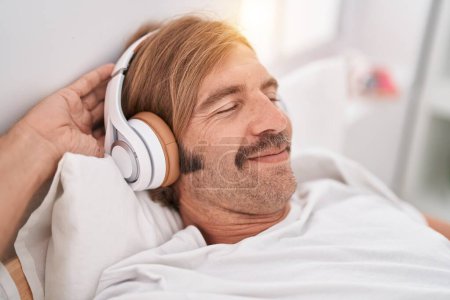 Téléchargez les photos : Jeune homme blond écoutant de la musique détendue sur le lit dans la chambre - en image libre de droit