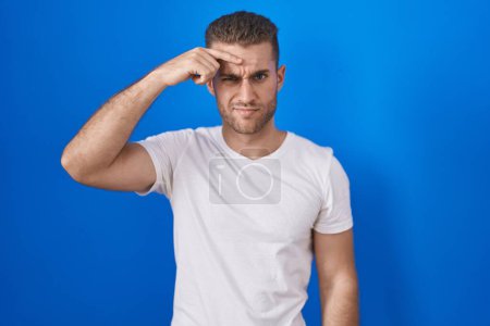 Foto de Young caucasian man standing over blue background pointing unhappy to pimple on forehead, ugly infection of blackhead. acne and skin problem - Imagen libre de derechos