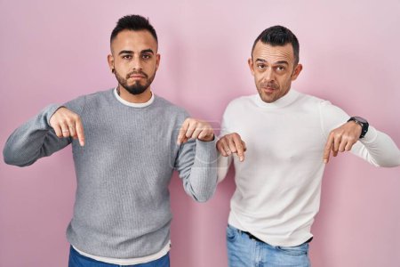 Photo for Homosexual couple standing over pink background pointing down looking sad and upset, indicating direction with fingers, unhappy and depressed. - Royalty Free Image