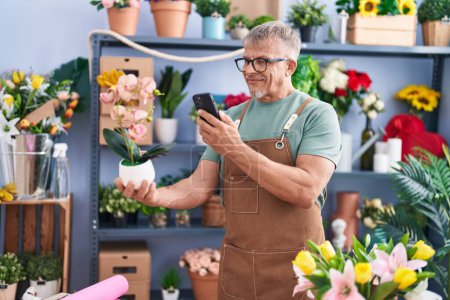 Foto de Middle age grey-haired man florist make photo to plant by smartphone at flower shop - Imagen libre de derechos
