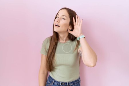 Téléchargez les photos : Beautiful brunette woman standing over pink background smiling with hand over ear listening an hearing to rumor or gossip. deafness concept. - en image libre de droit
