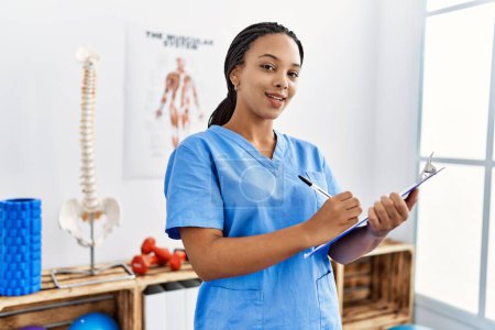 Téléchargez les photos : Young african american woman wearing physio therapist uniform writing on clipboard at clinic - en image libre de droit