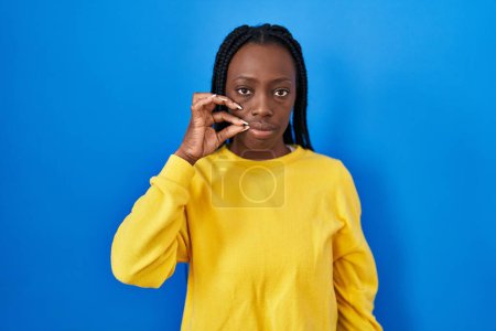Photo for Beautiful black woman standing over blue background mouth and lips shut as zip with fingers. secret and silent, taboo talking - Royalty Free Image