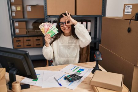 Téléchargez les photos : Young hispanic woman working at small business ecommerce holding money smiling happy doing ok sign with hand on eye looking through fingers - en image libre de droit