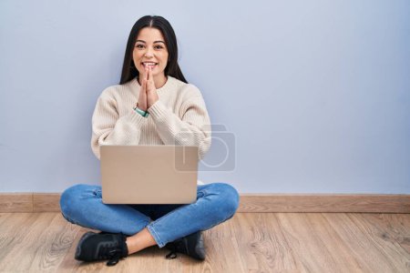 Téléchargez les photos : Jeune femme utilisant un ordinateur portable assis sur le sol à la maison priant avec les mains ensemble demandant pardon souriant confiant. - en image libre de droit