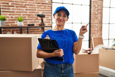 Photo for Middle age brunette woman wearing delivery uniform at house moving smiling happy pointing with hand and finger to the side - Royalty Free Image