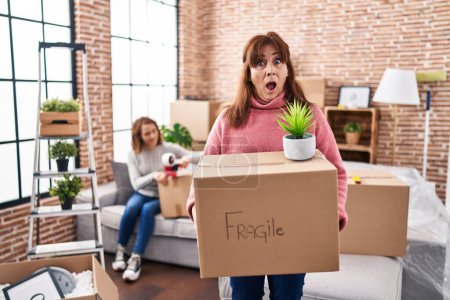 Foto de Mother and daughter moving to a new home holding cardboard box afraid and shocked with surprise and amazed expression, fear and excited face. - Imagen libre de derechos