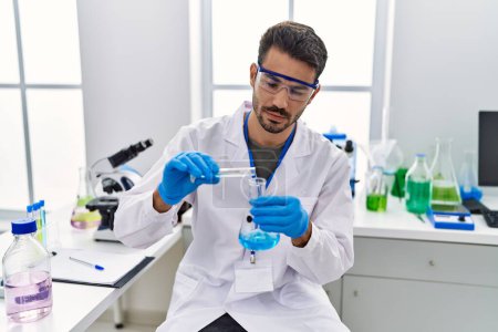 Foto de Joven hispano vistiendo uniforme científico vertiendo líquido en tubo de ensayo en laboratorio - Imagen libre de derechos