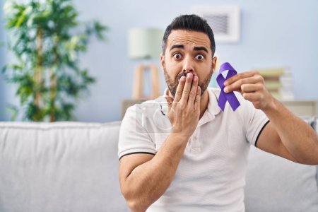 Photo for Young hispanic man with beard holding purple ribbon awareness covering mouth with hand, shocked and afraid for mistake. surprised expression - Royalty Free Image