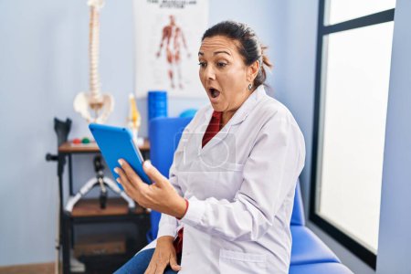 Foto de Middle age hispanic doctor woman working on online appointment scared and amazed with open mouth for surprise, disbelief face - Imagen libre de derechos