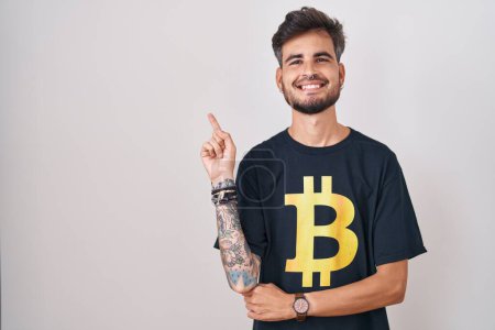 Photo for Young hispanic man with tattoos wearing bitcoin t shirt with a big smile on face, pointing with hand finger to the side looking at the camera. - Royalty Free Image