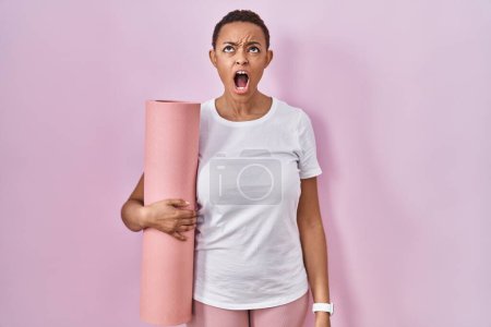 Photo for Beautiful african american woman holding yoga mat angry and mad screaming frustrated and furious, shouting with anger. rage and aggressive concept. - Royalty Free Image