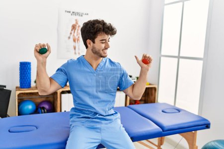Foto de Joven hombre hispano usando uniforme de fisioterapeuta usando bola anti estrés en la clínica - Imagen libre de derechos