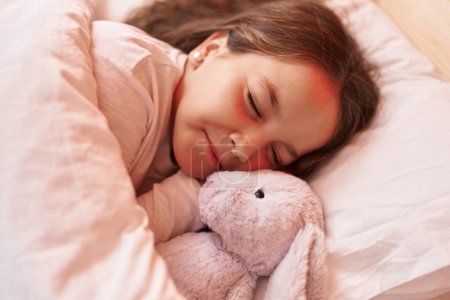 Foto de Adorable hispanic girl hugging rabbit doll lying on bed sleeping at bedroom - Imagen libre de derechos