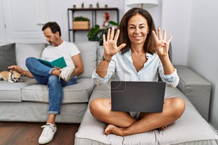 Foto de Pareja hispana de mediana edad en casa, mujer usando laptop mostrando y señalando con los dedos el número nueve mientras sonríe confiada y feliz. - Imagen libre de derechos