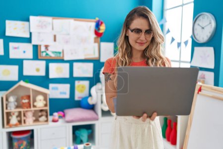 Téléchargez les photos : Jeune enseignante blonde utilisant un ordinateur portable debout à la maternelle - en image libre de droit