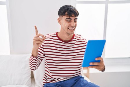 Téléchargez les photos : Jeune homme hispanique utilisant touchpad assis sur le lit souriant heureux pointant avec la main et le doigt sur le côté - en image libre de droit