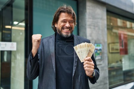 Guapo hombre de mediana edad sosteniendo billetes de 100 coronas danesas gritando orgulloso, celebrando la victoria y el éxito muy emocionado con el brazo levantado 