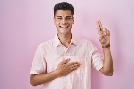 Foto de Joven hombre hispano de pie sobre fondo rosa sonriendo jurando con la mano en el pecho y los dedos hacia arriba, haciendo una promesa de lealtad juramento - Imagen libre de derechos