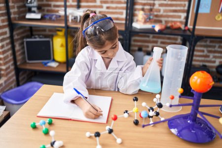 Foto de Adorable hispanic girl student holding test tube writing on notebook at laboratory classroom - Imagen libre de derechos