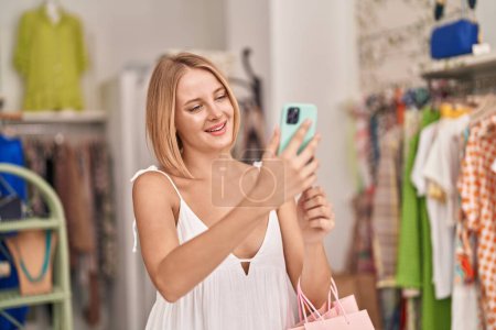 Téléchargez les photos : Jeune femme blonde client souriant confiant en utilisant un smartphone au magasin de vêtements - en image libre de droit