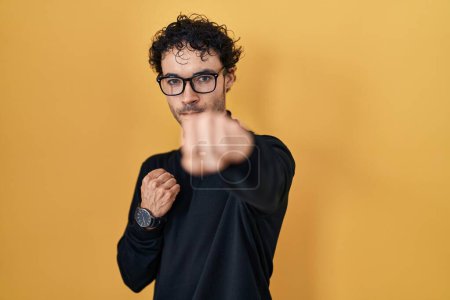 Foto de Hispanic man standing over yellow background punching fist to fight, aggressive and angry attack, threat and violence - Imagen libre de derechos