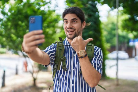 Photo for Hispanic man with long hair outdoors at the city doing video call pointing thumb up to the side smiling happy with open mouth - Royalty Free Image