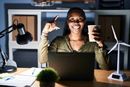 Photo for African woman working using computer laptop at night smiling and confident gesturing with hand doing small size sign with fingers looking and the camera. measure concept. - Royalty Free Image