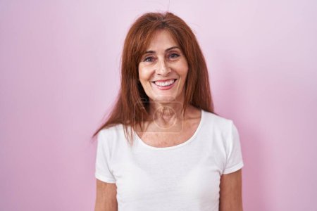 Foto de Mujer de mediana edad de pie sobre fondo rosa con una sonrisa feliz y fresca en la cara. persona afortunada. - Imagen libre de derechos