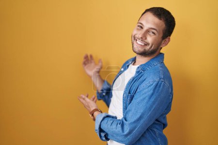 Foto de Hombre hispano de pie sobre fondo amarillo invitando a entrar sonriente natural con la mano abierta - Imagen libre de derechos