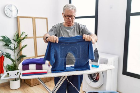 Photo for Senior caucasian man ironing holding burned iron shirt at laundry room skeptic and nervous, frowning upset because of problem. negative person. - Royalty Free Image