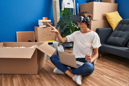 Foto de Young beautiful hispanic woman playing video game using virtual reality glasses sitting on floor at new home - Imagen libre de derechos