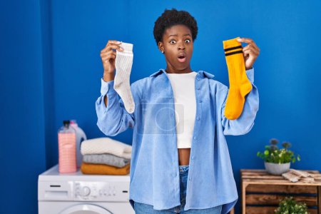Photo for African american woman holding clean andy dirty socks in shock face, looking skeptical and sarcastic, surprised with open mouth - Royalty Free Image