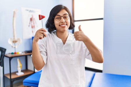 Photo for Young hispanic physiotherapist woman holding reflex hammer smiling happy and positive, thumb up doing excellent and approval sign - Royalty Free Image