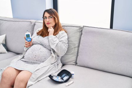 Photo for Pregnant woman using blood pressure monitor at home serious face thinking about question with hand on chin, thoughtful about confusing idea - Royalty Free Image