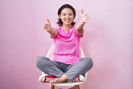Téléchargez les photos : Jeune femme hispanique assise sur une chaise sur fond rose regardant la caméra souriant à bras ouverts pour un câlin. expression joyeuse embrassant le bonheur. - en image libre de droit