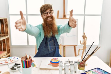 Foto de Pelirrojo con barba larga pintando tazón de barro en el estudio de arte mirando a la cámara sonriendo con los brazos abiertos para el abrazo. expresión alegre abrazando la felicidad. - Imagen libre de derechos