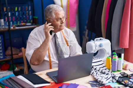 Téléchargez les photos : Homme moyen âge cheveux gris tailleur parler sur smartphone en utilisant un ordinateur portable au magasin de tailleur - en image libre de droit