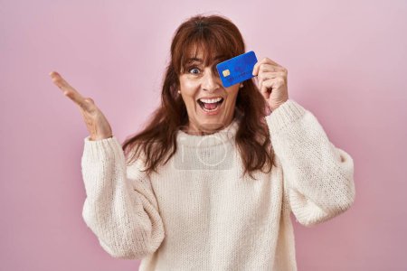 Photo for Middle age hispanic woman holding credit card covering eye celebrating victory with happy smile and winner expression with raised hands - Royalty Free Image