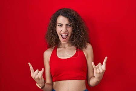 Téléchargez les photos : Femme hispanique avec les cheveux bouclés debout sur fond rouge criant avec une expression folle faire symbole rock avec les mains en l'air. star de la musique. concept de musique lourde. - en image libre de droit