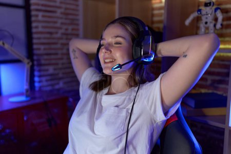 Foto de Joven mujer caucásica streamer sonriendo confiado relajado con las manos en la cabeza en la sala de juegos - Imagen libre de derechos