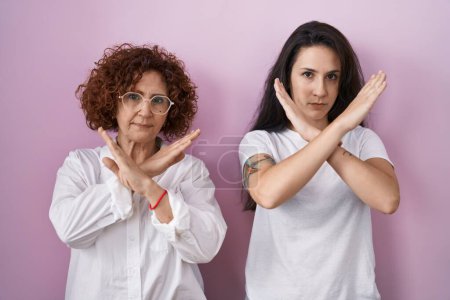 Foto de Madre e hija hispanas vistiendo camiseta blanca casual sobre expresión de rechazo de fondo rosa cruzando brazos haciendo signo negativo, cara enojada - Imagen libre de derechos